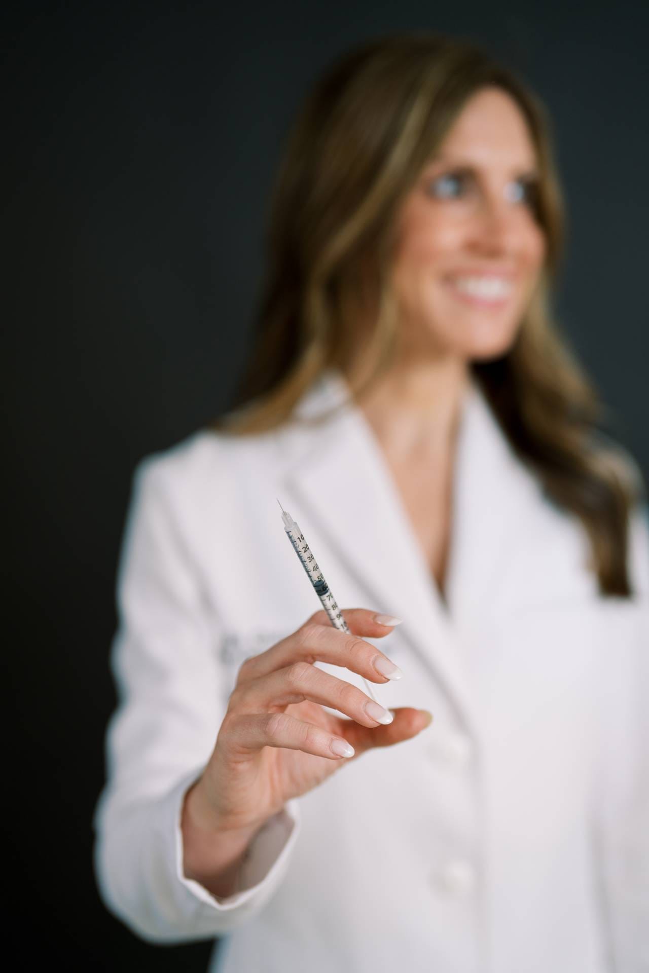A smiling healthcare provider in a white coat holds up a syringe in preparation for treatment