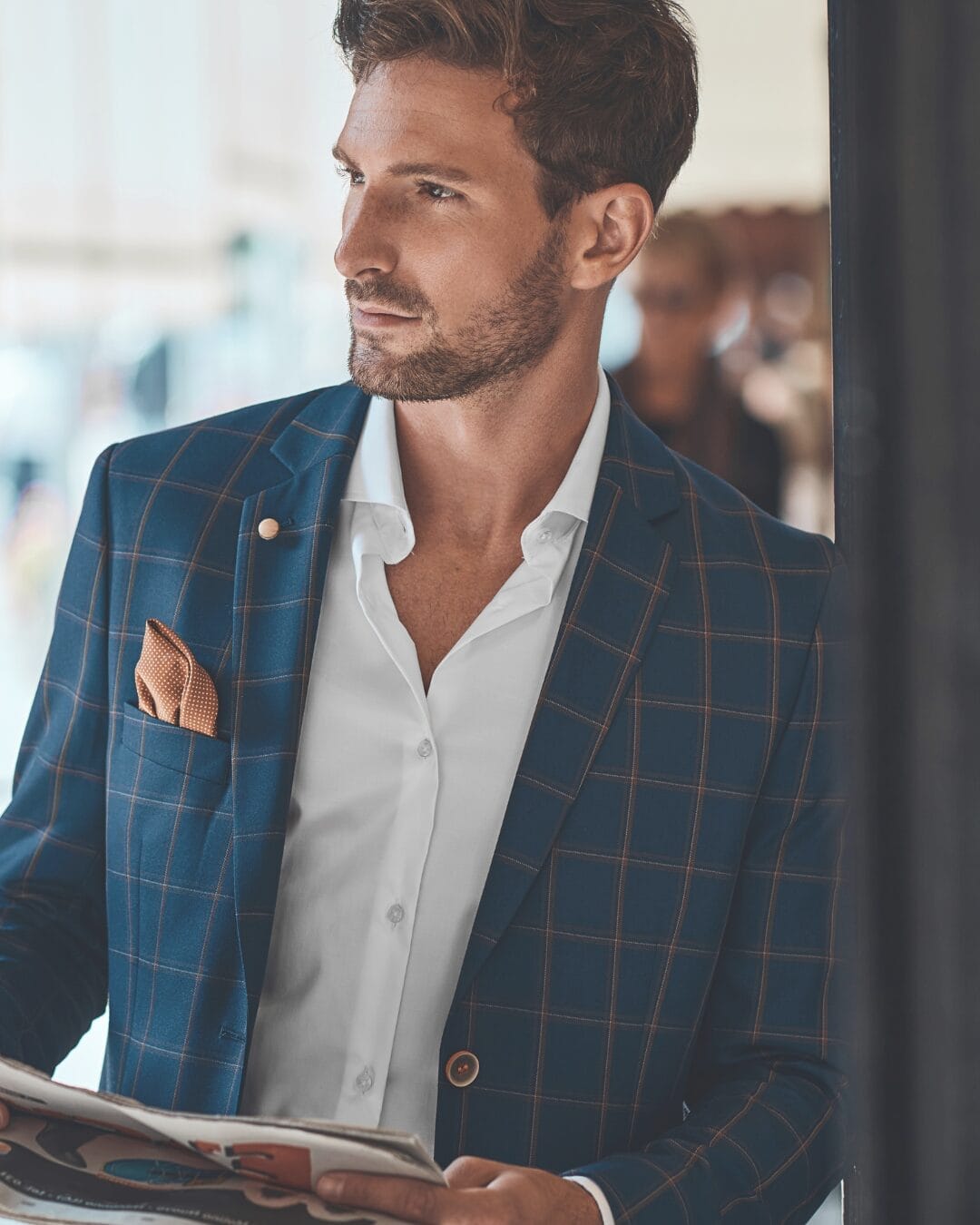 A man in a stylish blue plaid blazer with a pocket square looks off to the side while holding a newspaper